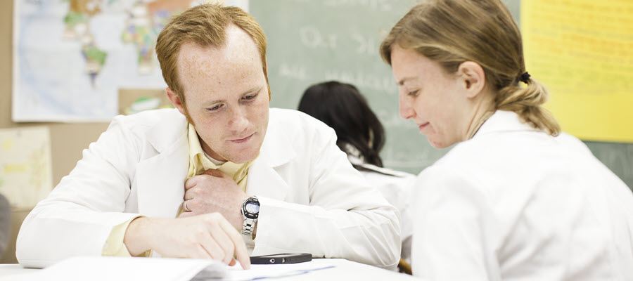 two students wearing white lab coats seated at a desk reviewing materials
