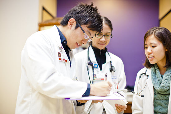 three physician students in white lab coats reviewing information on a clipboard